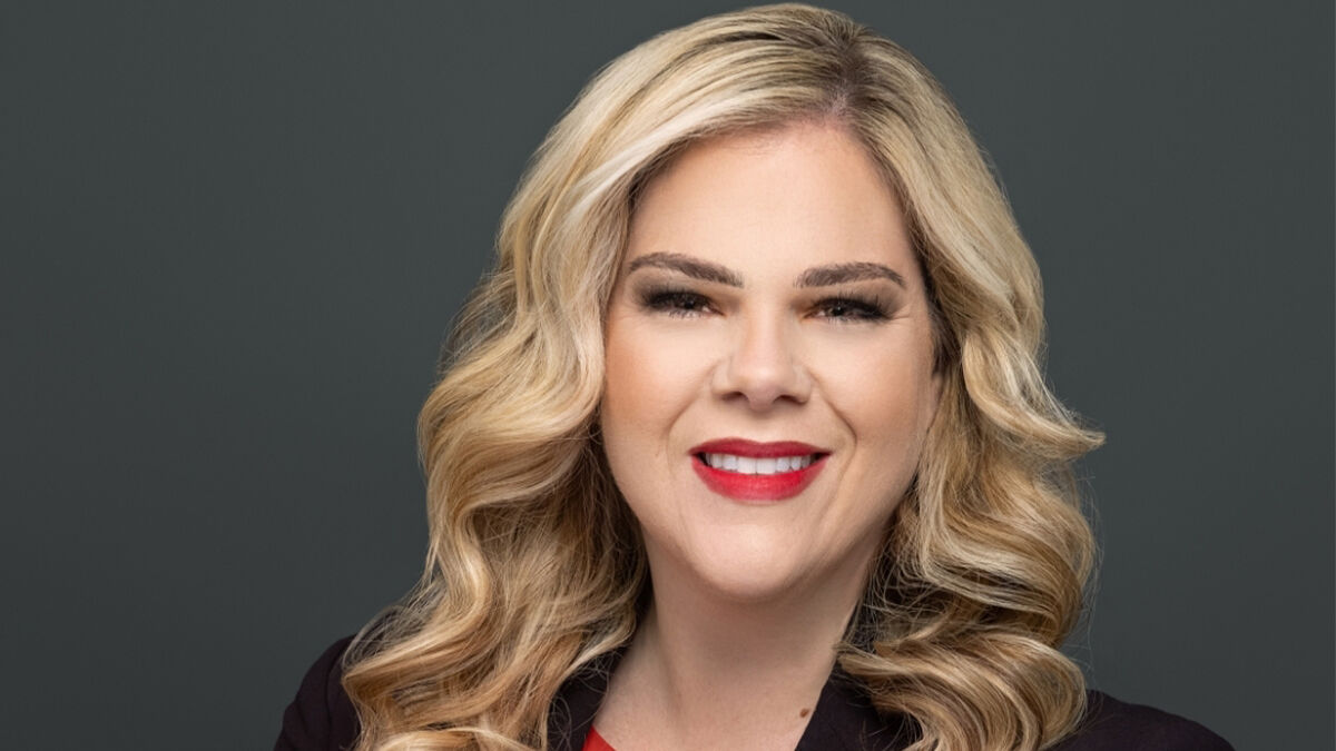 A person with long wavy blonde hair, wearing a black blazer and a red top, smiling in front of a plain dark background.