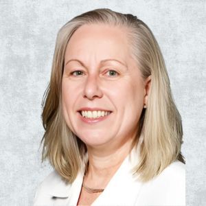 Portrait of a person with shoulder-length light hair and a white jacket, smiling against a gray textured background.