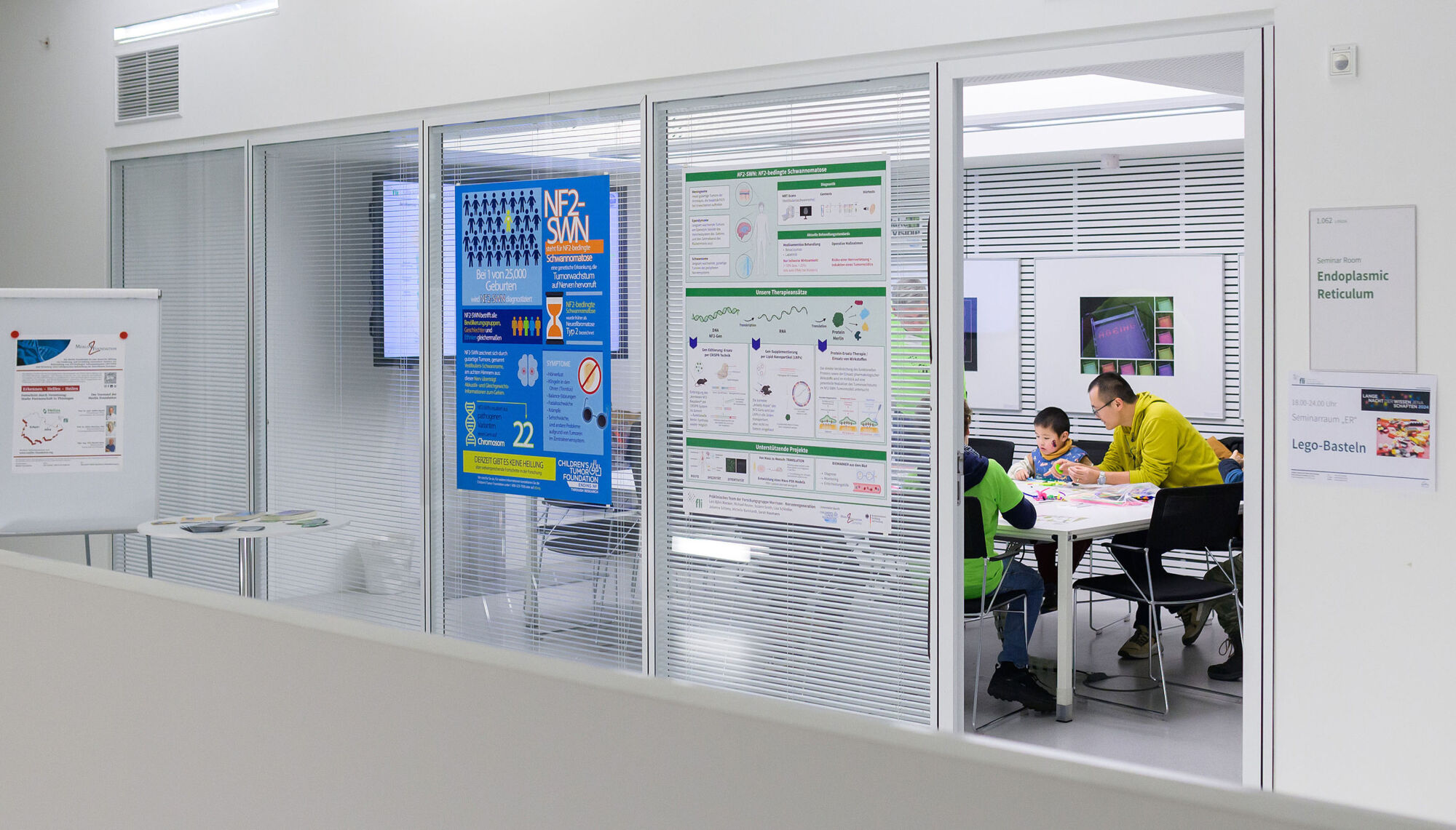 People sitting at a table in a glass-walled meeting room, with posters and informational signs on the walls and doors.