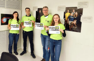 Four people wearing green shirts hold "Make NF Visible" signs, standing in a room with photos and text on the walls.