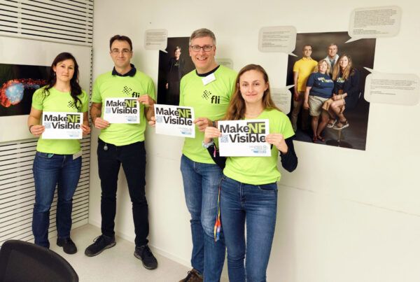 Four people wearing green shirts hold "Make NF Visible" signs, standing in a room with photos and text on the walls.