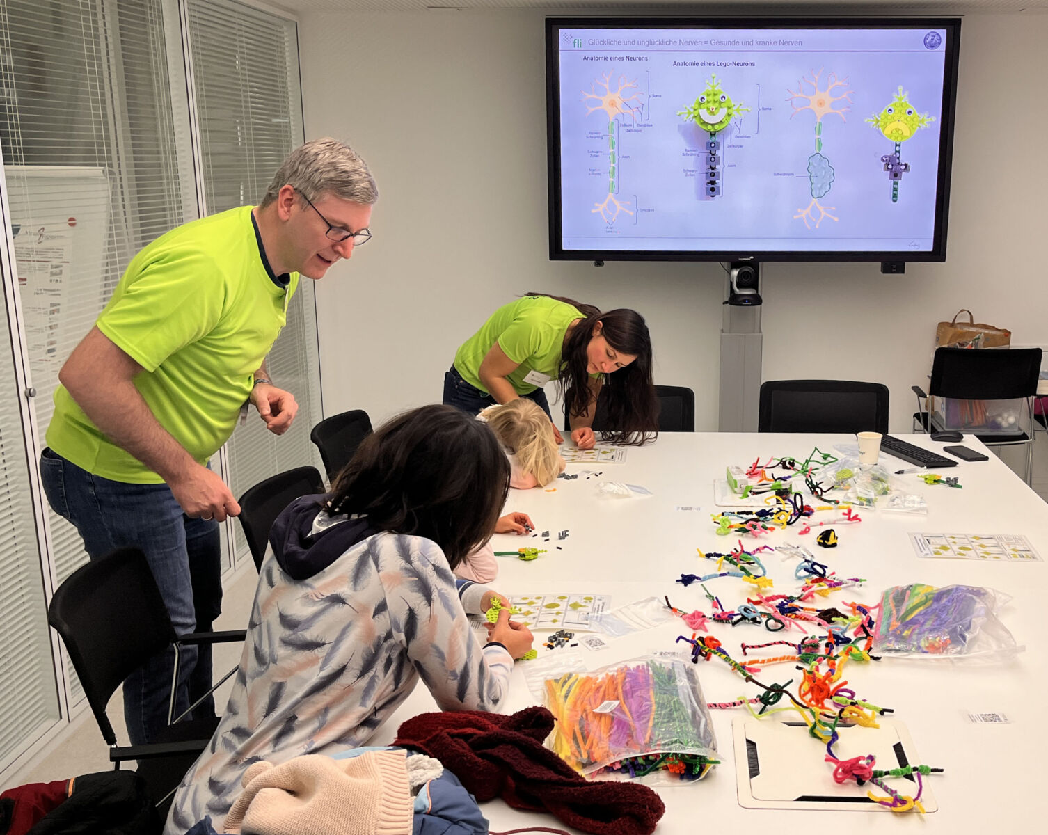 Two adults and children work on craft projects at a table with colorful materials. A screen displays diagrams in the background.