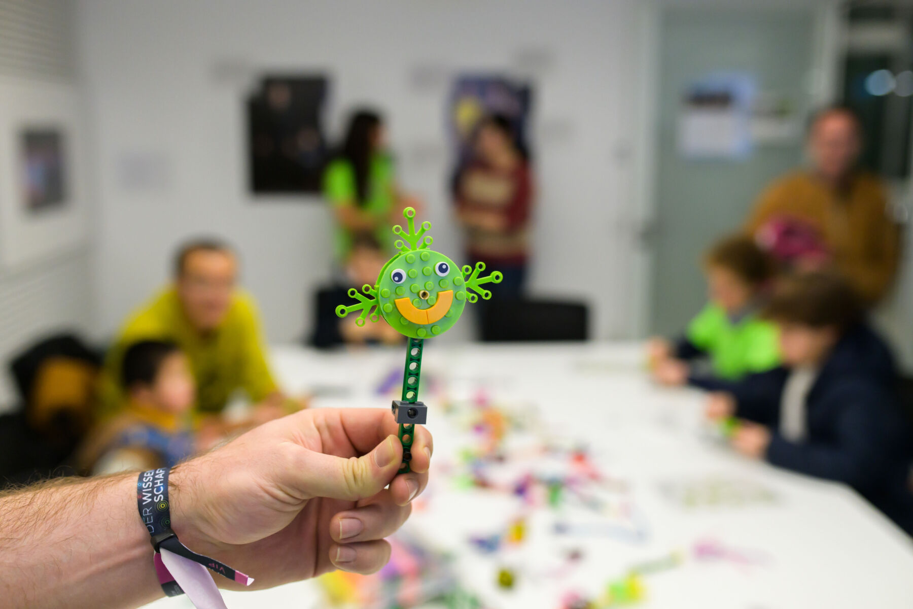 A hand holds a green smiling craft stick figure with a workshop in the background where people are seated at a table doing crafts.