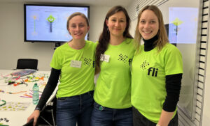 Three people wearing matching neon green shirts with the "fli" logo, standing in a conference room with a presentation screen in the background.