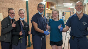 A group of four healthcare professionals and a patient stand in a hallway, all giving a thumbs-up gesture.