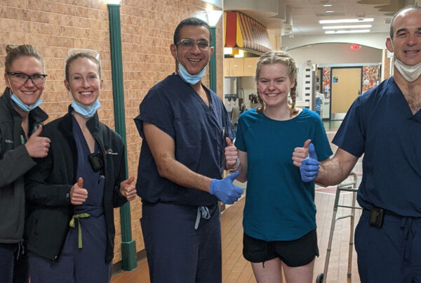 A group of four healthcare professionals and a patient stand in a hallway, all giving a thumbs-up gesture.