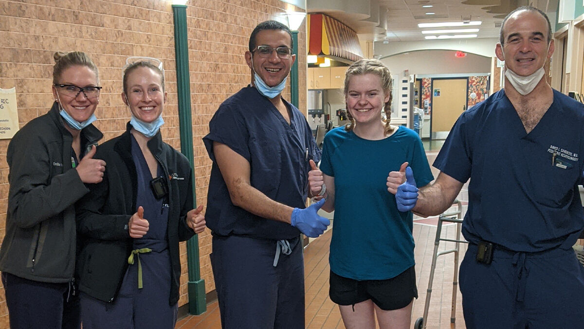 A group of four healthcare professionals and a patient stand in a hallway, all giving a thumbs-up gesture.