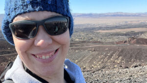 Person wearing sunglasses and a blue beanie smiling at the camera with a rocky landscape and distant mountains in the background.