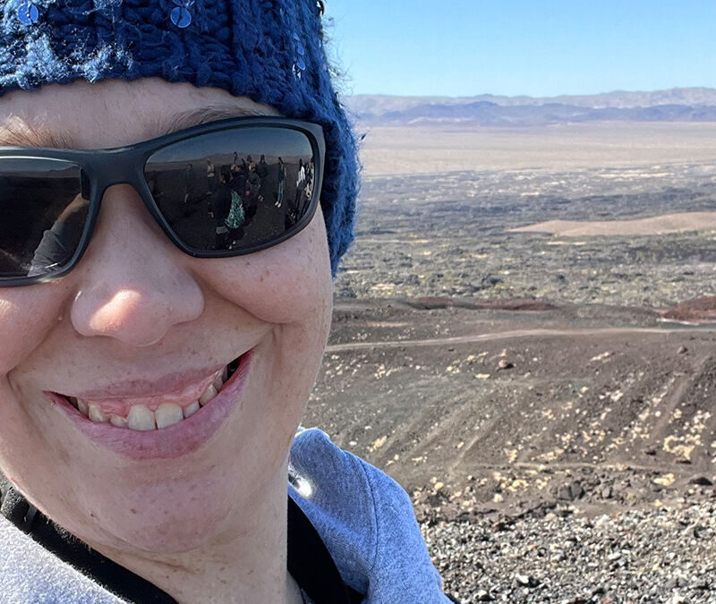 Person wearing sunglasses and a blue beanie smiling at the camera with a rocky landscape and distant mountains in the background.