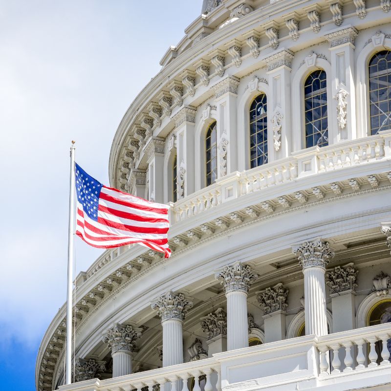 A U.S. flag waves in front of the United States Capitol building's white exterior, featuring columns and arched windows, under a partly cloudy sky.