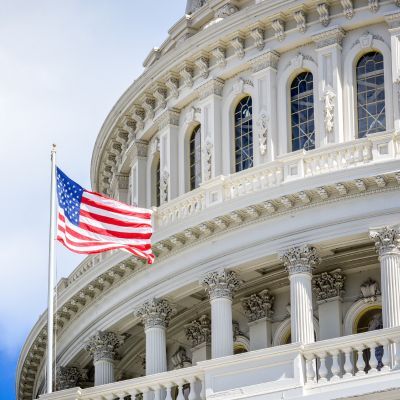 The United States Capitol dome with an American flag waving in front, set against a partly cloudy sky.