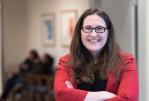 A woman with long brown hair and glasses smiles while crossing her arms. She is wearing a red blazer. The background shows blurred people sitting and framed artwork on the wall.