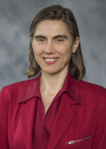 A person with shoulder-length hair is wearing a red blazer over a red blouse, smiling at the camera against a gray background.