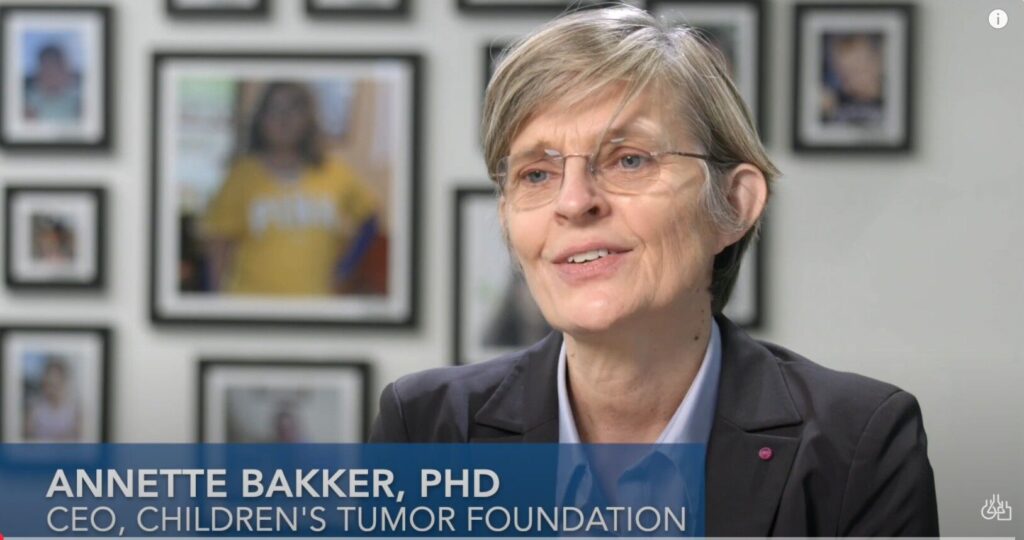 A person with short hair and glasses speaks in an interview. Several framed photos are visible in the blurred background. Text reads: "Annette Bakker, PhD, CEO, Children's Tumor Foundation.