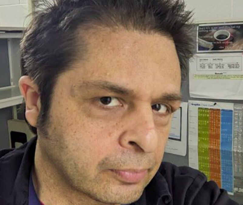 Person with short dark hair in an office setting, standing in front of a cubicle wall with papers and a headset visible in the background.