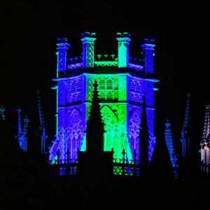 A castle tower illuminated in blue and green lights, surrounded by ornate spires, against a dark night sky.