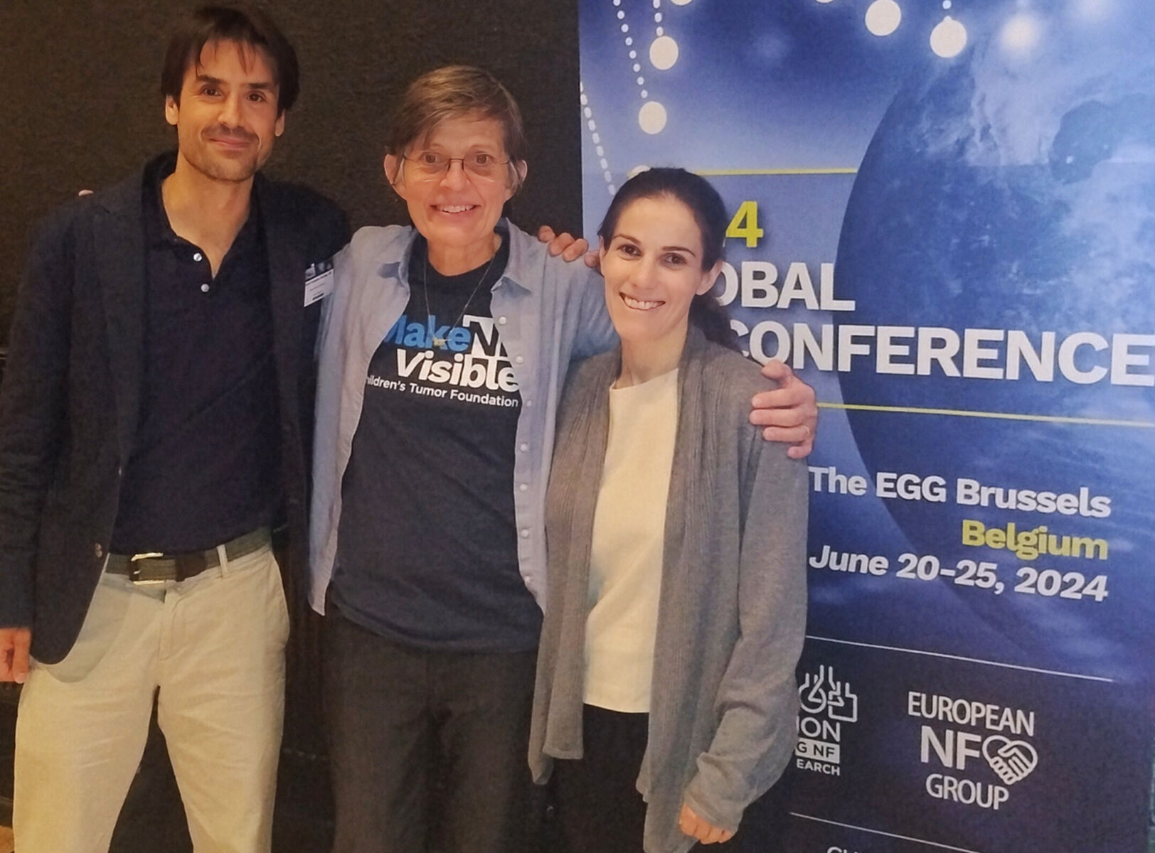 Three people smiling and posing in front of a banner for the 2024 Global Conference, held in Brussels from June 20-25, 2024.