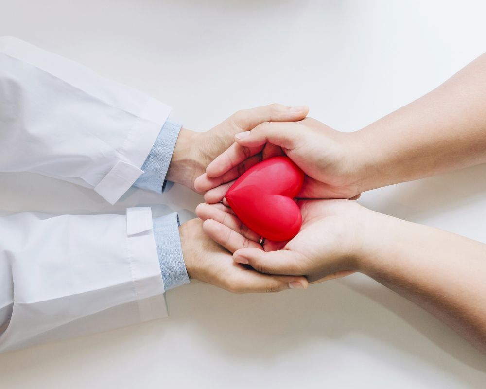 Two pairs of hands hold a red heart-shaped object together. One person wears a white long-sleeved garment, possibly a lab coat, and the other has bare arms.